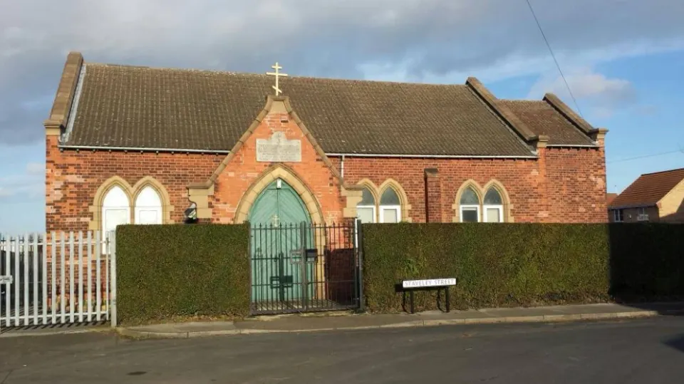 The church from Staveley Street: a red-brick building behind a hedge, with arched windows, a green door and a gold three-bar cross on the gable
